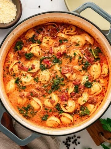 Tuscan Tortellini soup with ground meat and kale in a creamy red tomato-based sauce, served in a Dutch oven with a side of grated cheese.
